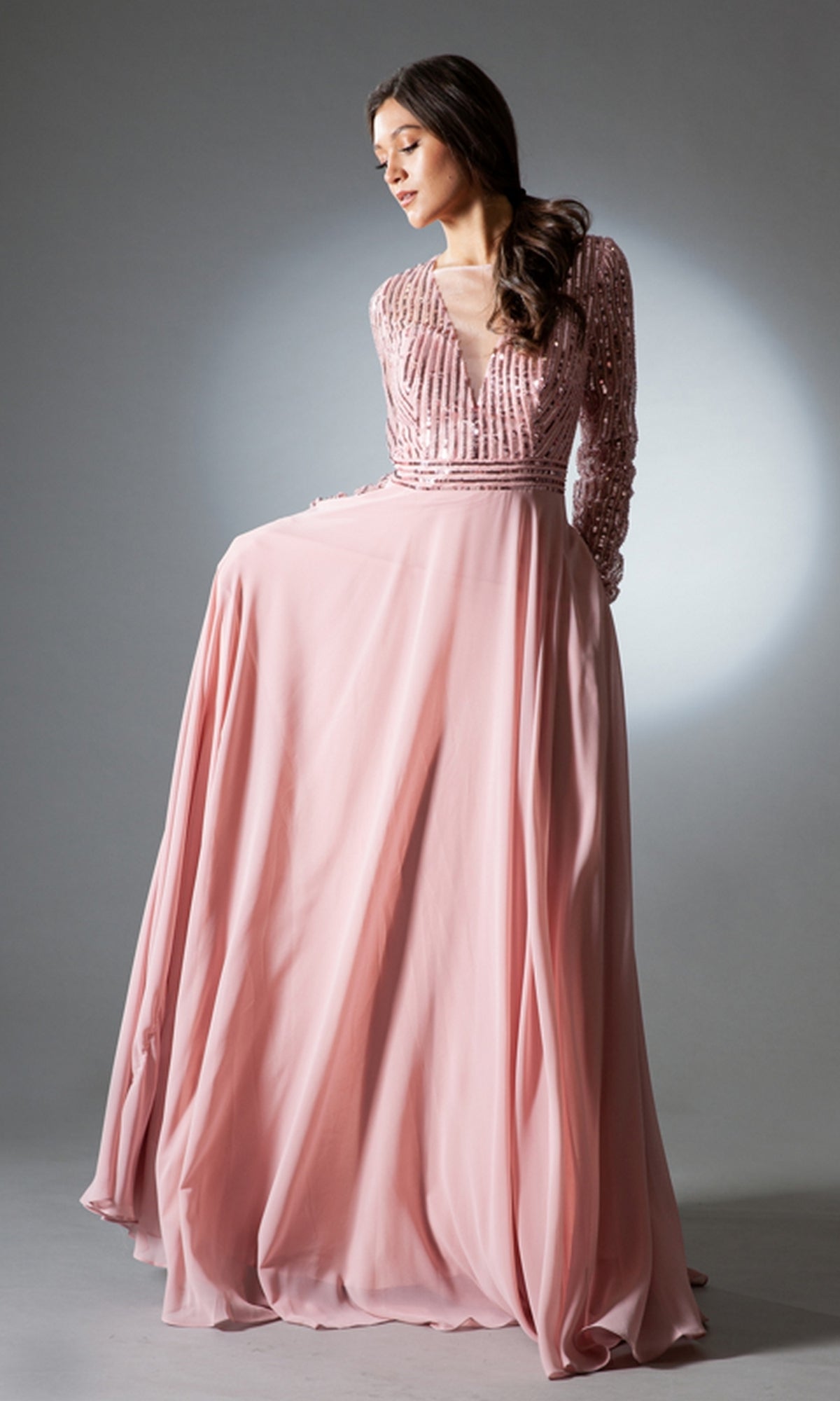 A woman stands against a gray background wearing the Long-Sleeve Long Modest Prom Dress 7036—a flowing pink evening gown with sequin bodice and long sleeves.